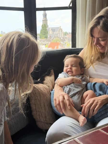 A heartwarming family moment unfolds indoors, with a mother tenderly holding a smiling baby while an older child looks on, all bathed in bright daylight